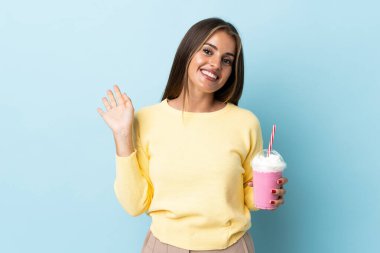 Çilekli milkshake 'li genç Uruguaylı kadın mavi arka planda izole edilmiş mutlu bir ifadeyle selamlıyor.