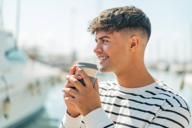 Dışardaki Latin kökenli genç adam elinde kahve tutuyor.