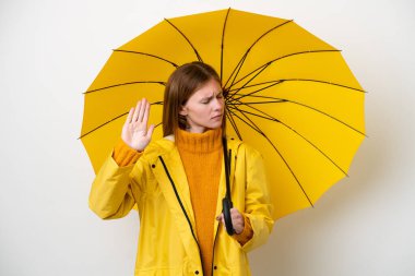 Yağmura dayanıklı ceketi ve şemsiyesi olan genç bir İngiliz kadın beyaz arka planda izole edilmiş, durdurma hareketi yapıyor ve hayal kırıklığına uğruyor