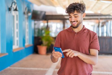 Dışarıdaki yakışıklı genç Arap adam cep telefonuyla mesaj ya da e-posta yolluyor.