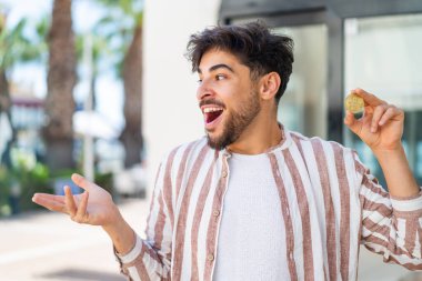 Yakışıklı bir Arap açık havada Bitcoin tutuyor ve yüzünde sürpriz bir ifade var.