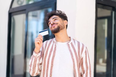 Açık havada elinde kredi kartıyla dolaşan yakışıklı bir Arap.