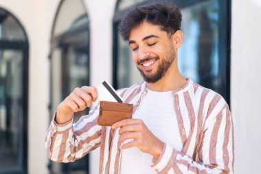 Açık havada yakışıklı bir Arap 'ın elinde cüzdan ve mutlu bir ifadeyle kredi kartı.