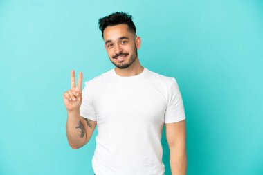 Mavi arka planda izole edilmiş genç beyaz adam gülümsüyor ve zafer işareti gösteriyor.