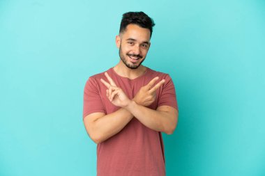 Mavi arka planda izole edilmiş genç beyaz adam gülümsüyor ve zafer işareti gösteriyor.