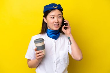 Çinli kadın hostes, sarı arka planda izole edilmiş. Elinde kahve ve cep telefonu var.