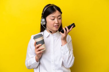 Tele pazarlamacı Çinli bir kadın sarı arka planda izole edilmiş bir kulaklıkla çalışıyor. Elinde kahve ve cep telefonu var.