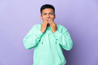 Genç Ekvadorlu adam mor arka planda izole edilmiş mutlu ve gülümseyen elleri ile ağzını kapatıyor.