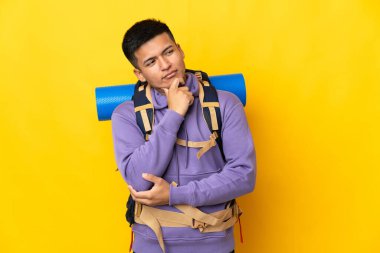 Sarı arka planda izole edilmiş büyük bir sırt çantası olan genç dağcı yana bakıyor ve gülümsüyor.