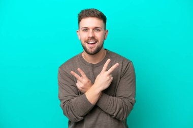 Mavi arka planda izole edilmiş genç beyaz adam gülümsüyor ve zafer işareti gösteriyor.