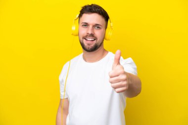 Genç beyaz adam sarı arka planda izole edilmiş müzik dinliyor ve başparmağını kaldırıyor.