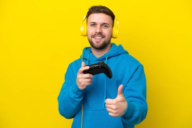 Sarı arka planda izole edilmiş bir bilgisayar oyunu kontrolörüyle oynayan genç beyaz bir adam işaret veriyor.