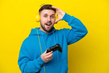 Sarı arka planda izole edilmiş bir bilgisayar oyunu kontrolörüyle oynayan genç beyaz adam yan tarafa bakarken sürpriz jest yapıyor.