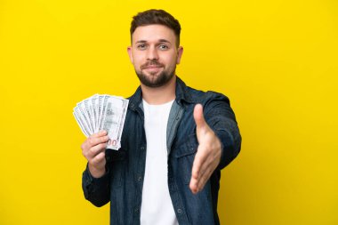 Kafkasyalı genç adam sarı arka planda izole edilmiş bir sürü para alıyor ve iyi bir anlaşma için el sıkışıyor.