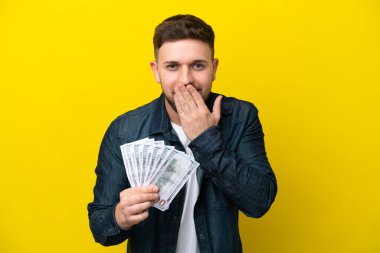 Sarı arka planda izole edilmiş bir sürü para alan beyaz bir genç adam mutlu ve gülümseyen bir ağızla elini kapatıyor.
