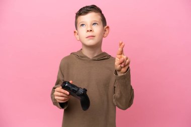 Küçük beyaz çocuk pembe arka planda izole edilmiş bir bilgisayar oyunu kontrolörüyle oynuyor. Parmaklarını çapraz yapıp en iyisini diliyor.