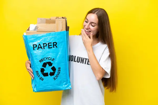 若い白人女性は 驚きと衝撃的な表情で黄色の背景に隔離されたリサイクルするために紙でいっぱいのリサイクルバッグを保持しています — ストック写真