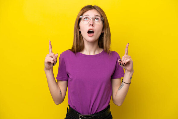 Молодая английская женщина, изолированная на жёлтом фоне, удивлена и указывает вверх