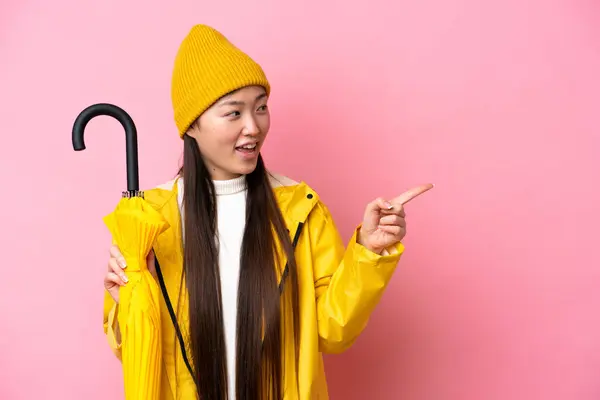 Young Chinese woman with rainproof coat and umbrella isolated on pink background pointing finger to the side and presenting a product