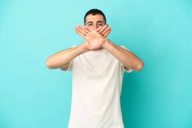 Genç, beyaz, mavi arka planda izole edilmiş genç bir adam bir eylemi durdurmak için eliyle dur hareketi yapıyor.