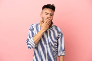 Pembe arka planda izole edilmiş genç beyaz adam esniyor ve ağzını eliyle açık tutuyor.