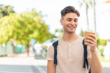 Genç yakışıklı bir adam açık havada elinde kahveyle neşeli bir ifade ile geziyor.