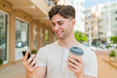 Genç yakışıklı adam cep telefonu kullanıyor ve elinde mutlu bir ifadeyle kahve tutuyor.