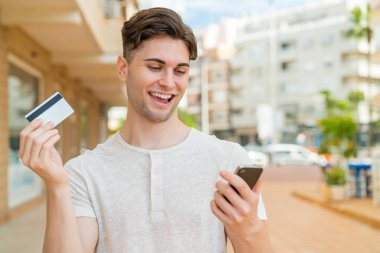 Genç yakışıklı adam cep telefonuyla kredi kartıyla alışveriş yapıyor.