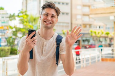 Dışarıda cep telefonu kullanan genç yakışıklı adam mutlu bir ifadeyle selamlıyor.