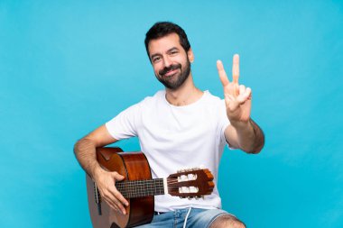 Mavi arka planda gitar çalan genç adam gülümsüyor ve zafer işareti gösteriyor.