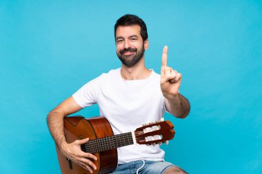 İzole edilmiş mavi arka planda gitar çalan genç bir adam parmağını kaldırıyor ve gösteriyor.