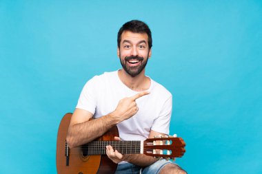 İzole edilmiş mavi arka planda gitar çalan genç adam şaşırmış ve işaret ediyor.