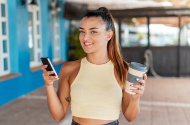 Açık havada cep telefonu kullanan ve elinde mutlu bir ifadeyle kahve tutan genç ve güzel bir kadın.