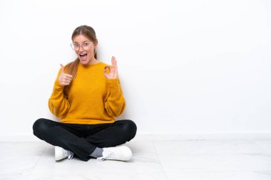 Beyaz arka planda izole bir şekilde yerde oturan beyaz tenli genç bir kadın tamam işareti ve baş parmak hareketi gösteriyor.