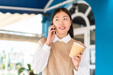 Dışarıdaki genç Çinli kadın cep telefonu kullanıyor ve elinde kahve tutuyor.