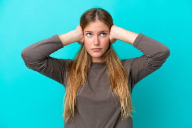 Mavi arka planda izole edilmiş genç sarışın kadın hayal kırıklığına uğramış ve kulaklarını kapatmış.
