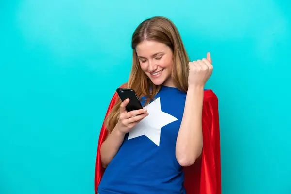 スーパーヒーローCaucasian女性隔離上の青の背景で電話で勝利位置 ストックフォト