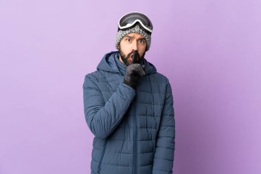 İzole edilmiş mor arka planda snowboard gözlüklü kayakçı parmağını ağzına koyan sessizlik işareti gösteriyor.