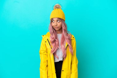 Pembe saçlı, yağmura dayanıklı bir ceket giyen, mavi arka planda izole edilmiş genç bir kadın.