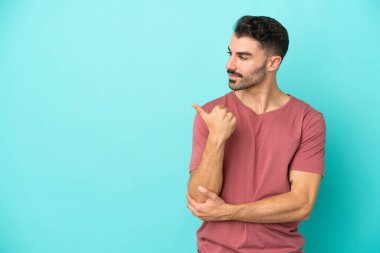 Mavi arka planda izole edilmiş genç beyaz adam bir ürünü sunmak için kenarı işaret ediyor.