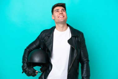 Motosiklet kasklı genç beyaz adam mavi arka planda izole edilmiş yukarı bakarken bir fikir düşünüyor.