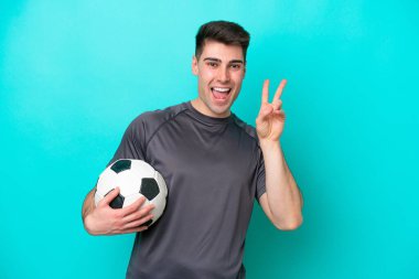 Kafkasyalı genç futbolcu mavi arka planda izole edilmiş gülümsüyor ve zafer işareti gösteriyor.