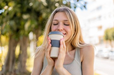 Sarışın bir kadın açık havada elinde kahveyle neşeli bir ifade ile bekliyor.