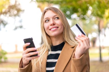 Genç, güzel, sarışın bir kadın dışarıda cep telefonuyla alışveriş yapıyor ve elinde şaşırmış bir ifadeyle bir kredi kartı tutuyor.