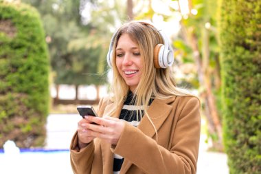 Açık havada müzik dinleyen güzel sarışın kadın cep telefonuyla mesaj yolluyor.