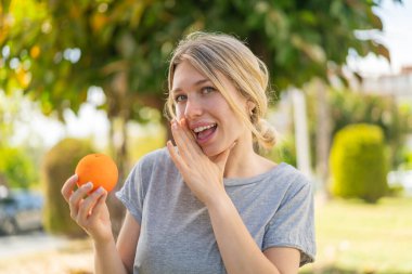 Açık havada elinde portakal tutan genç sarışın kadın bir şeyler fısıldıyor.