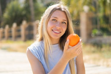 Açık havada genç sarışın bir kadın elinde mutlu bir ifadeyle bir portakal tutuyor.