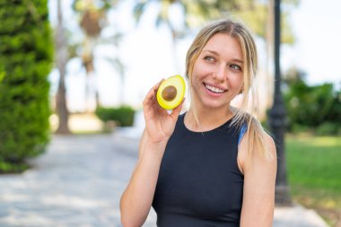 Açık havada elinde mutlu bir ifadeyle bir avokado tutan genç sarışın kadın.