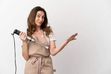 Orta yaşlı beyaz kadın el blenderi kullanıyor. Beyaz arka planda izole edilmiş. Gelmesi için ellerini yana uzatıyor.