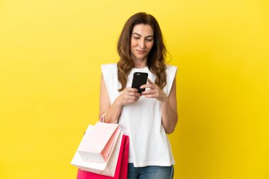Orta yaşlı beyaz kadın, sarı arka planda izole edilmiş. Elinde alışveriş poşetleri var ve bir arkadaşına cep telefonuyla bir mesaj yazıyor.